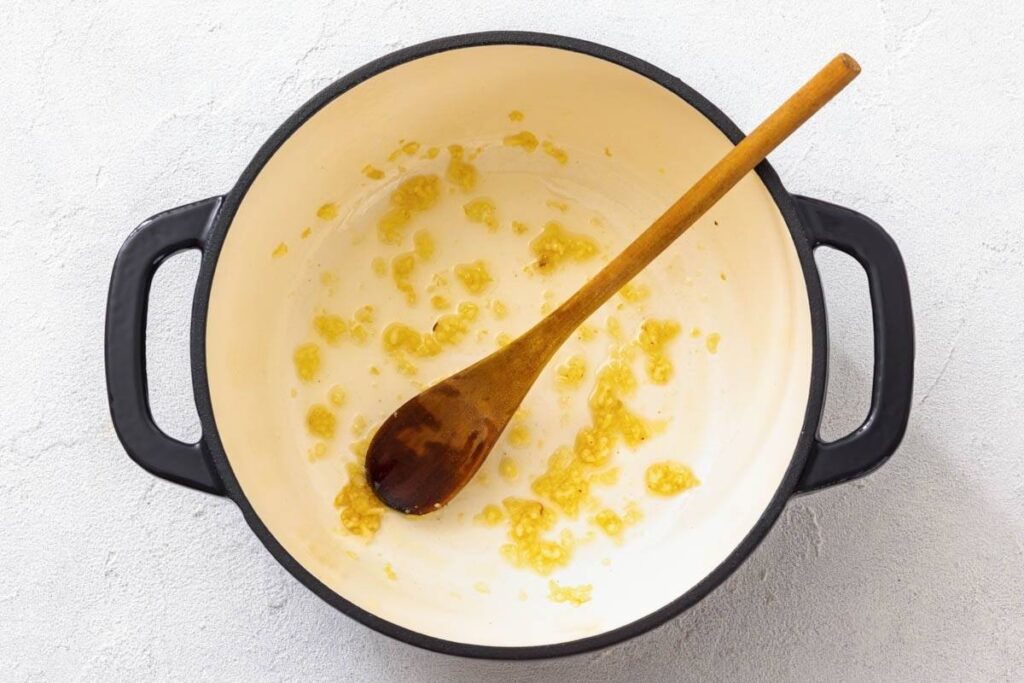 Chopped garlic is being sautéed in oil in a large white pot with a wooden spoon resting inside.