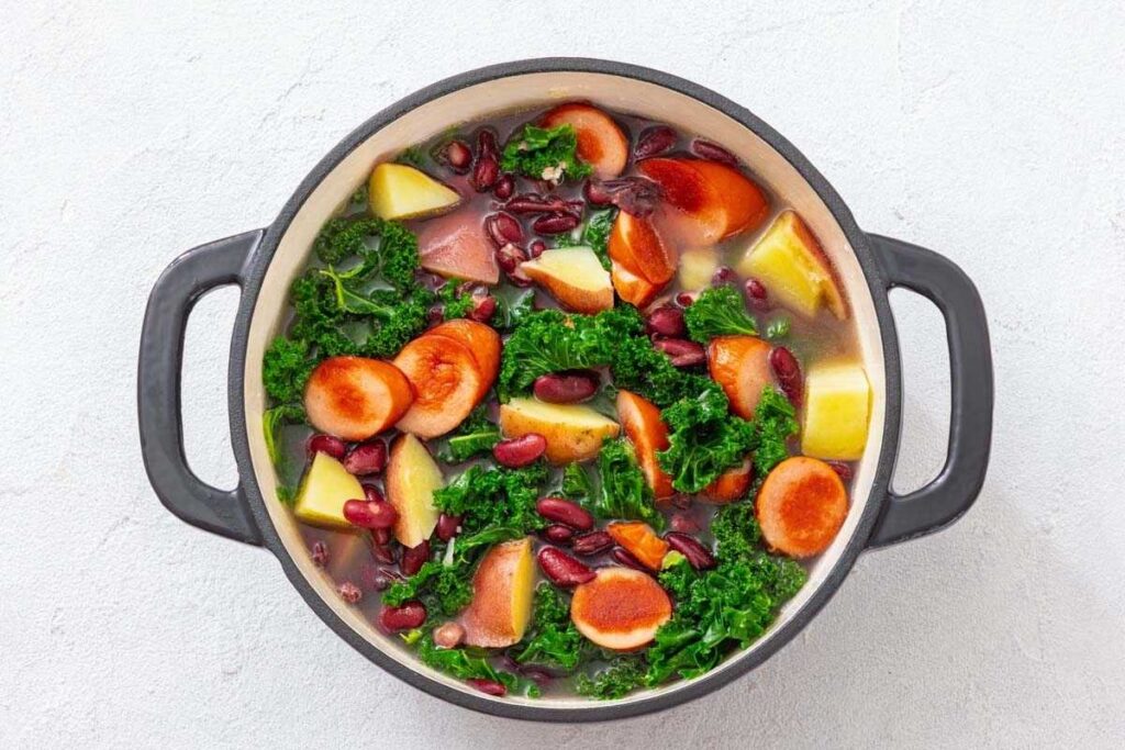 A pot of vegetable soup with sliced carrots, kidney beans, kale, and potato chunks in a light broth on a white background.