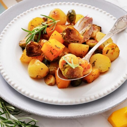 A white plate with roasted potatoes, Brussels sprouts, and butternut squash garnished with herbs, served with a spoon.