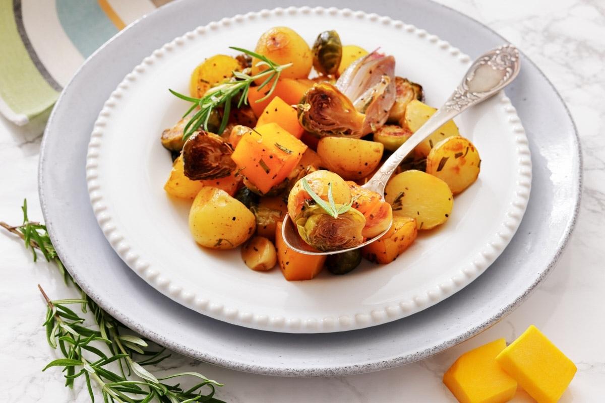 A white plate with roasted potatoes, Brussels sprouts, and butternut squash garnished with herbs, served with a spoon.