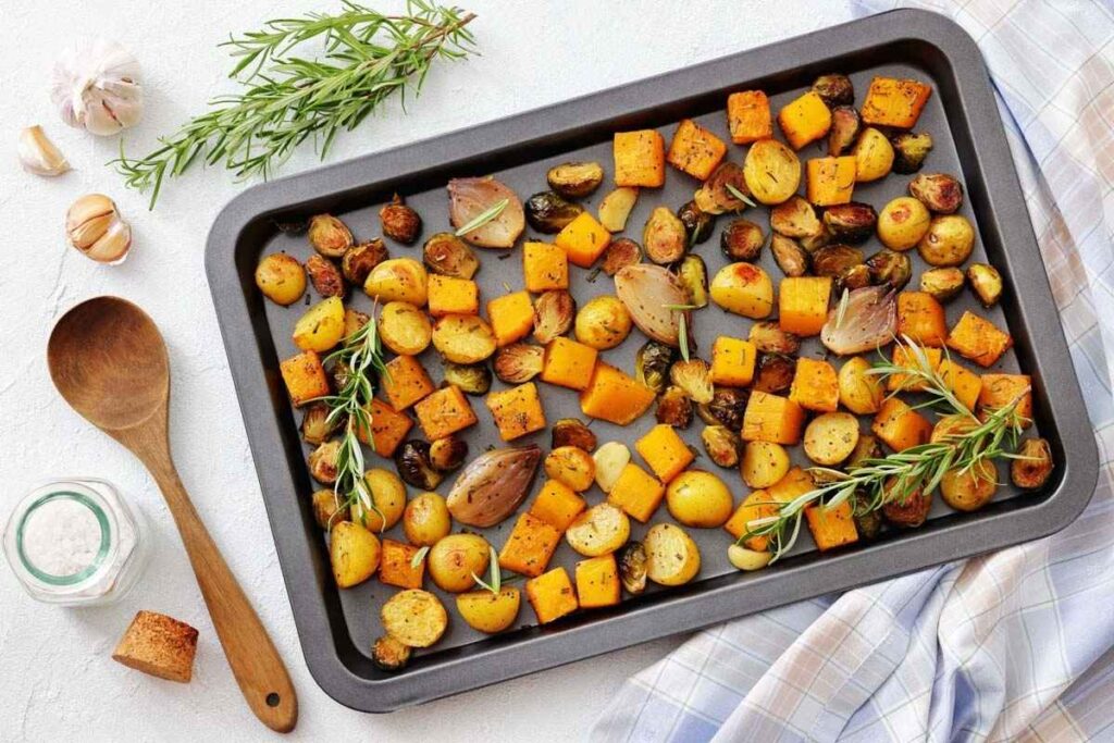 A baking tray with roasted vegetables including potatoes, brussels sprouts, shallots, and squash, garnished with rosemary. Garlic, salt, and a wooden spoon are nearby.