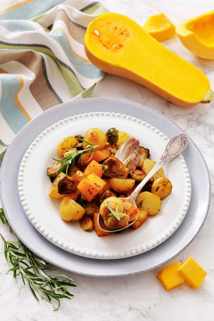 A plate of roasted vegetables, including potatoes and squash, garnished with herbs, sits on a white plate with a silver fork. A halved butternut squash is nearby.
