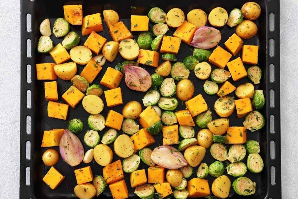 A baking tray with evenly spread pieces of yellow squash, potatoes, Brussels sprouts, and shallots, all seasoned and ready for roasting.