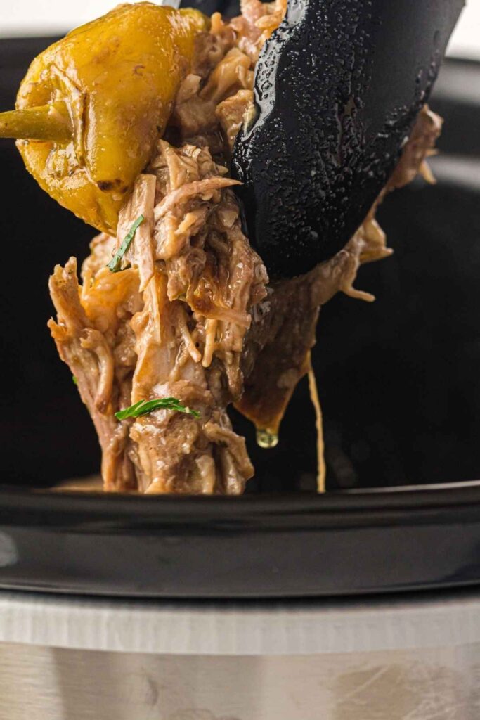 A close-up of shredded beef and a pepper being lifted from a slow cooker with black tongs.