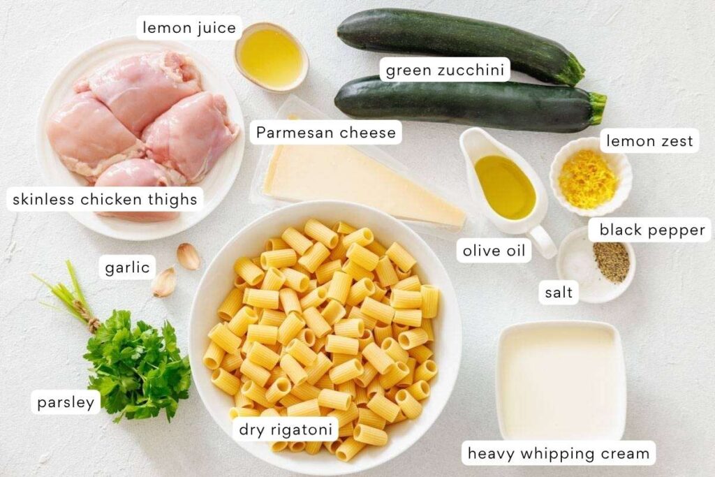 A top-down view of ingredients for a pasta dish including skinless chicken thighs, dry rigatoni, zucchini, parsley, parmesan, garlic, lemon, olive oil, salt, pepper, and heavy cream.