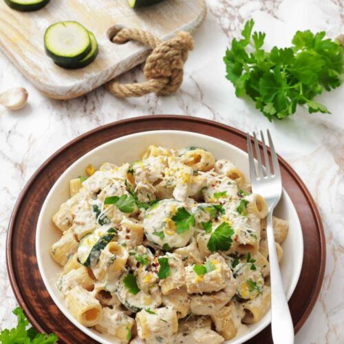 A bowl of rigatoni pasta with creamy sauce, zucchini slices, and parsley, placed on a marble surface beside a fork, fresh parsley, a lemon half, and a cutting board with zucchini.