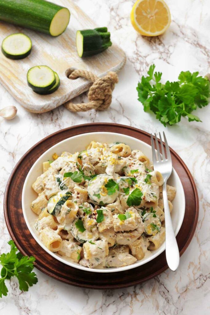 A bowl of rigatoni pasta with creamy sauce, zucchini slices, and parsley, placed on a marble surface beside a fork, fresh parsley, a lemon half, and a cutting board with zucchini.