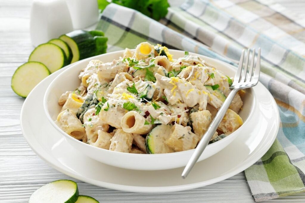 A white bowl of creamy rigatoni pasta with sliced zucchini, herbs, and lemon zest, served with a fork on a white plate, placed on a checkered cloth.