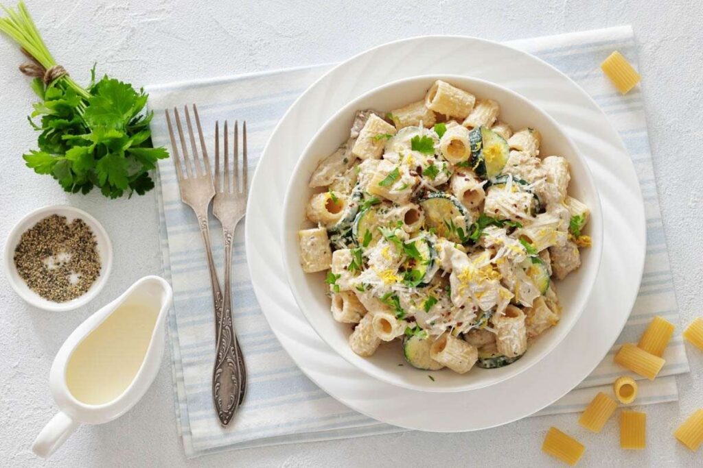 A plate of creamy pasta with herbs and vegetables sits on a white and blue napkin, accompanied by two forks, a small bowl of pepper, fresh parsley, and a sauce container.