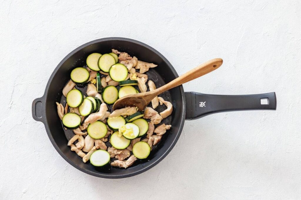 A frying pan with sliced zucchini and pieces of chicken being cooked, mixed with a wooden spoon, on a light surface.