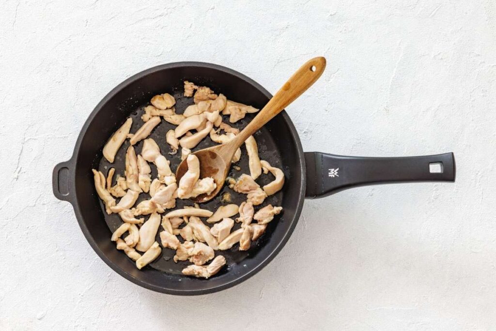 A frying pan with sliced pieces of chicken being cooked, stirred with a wooden spoon on a light-colored surface.