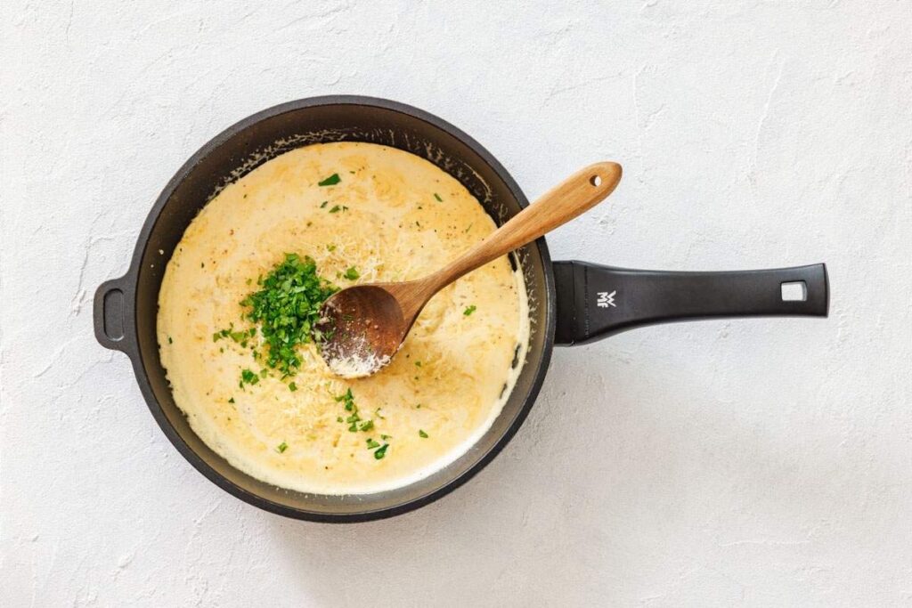 A black frying pan with a creamy sauce, grated cheese, and chopped parsley being mixed with a wooden spoon on a white surface.