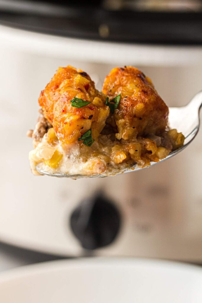 A close-up of a spoonful of tater tot casserole with ground meat, sauce, and herbs, held in front of a slow cooker.