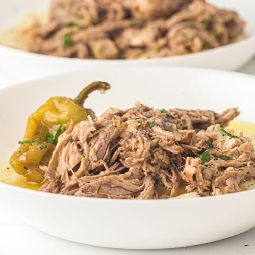 Shredded beef served over mashed potatoes with a whole pepper and garnished with herbs in a white bowl.