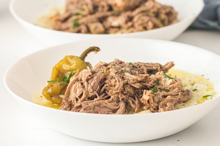 Shredded beef served over mashed potatoes with a whole pepper and garnished with herbs in a white bowl.