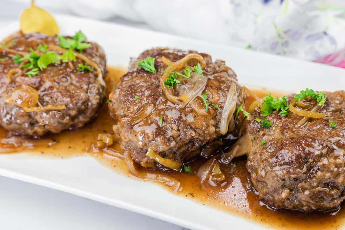 Three Mississippi Salisbury Steak covered in brown onion gravy, garnished with chopped parsley, served on a white rectangular plate.