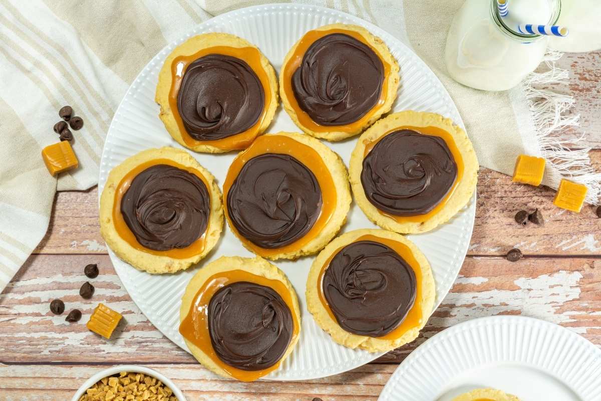 A white plate with seven cookies topped with caramel and a layer of swirled chocolate, placed on a wooden table with scattered caramel pieces and chocolate chips.