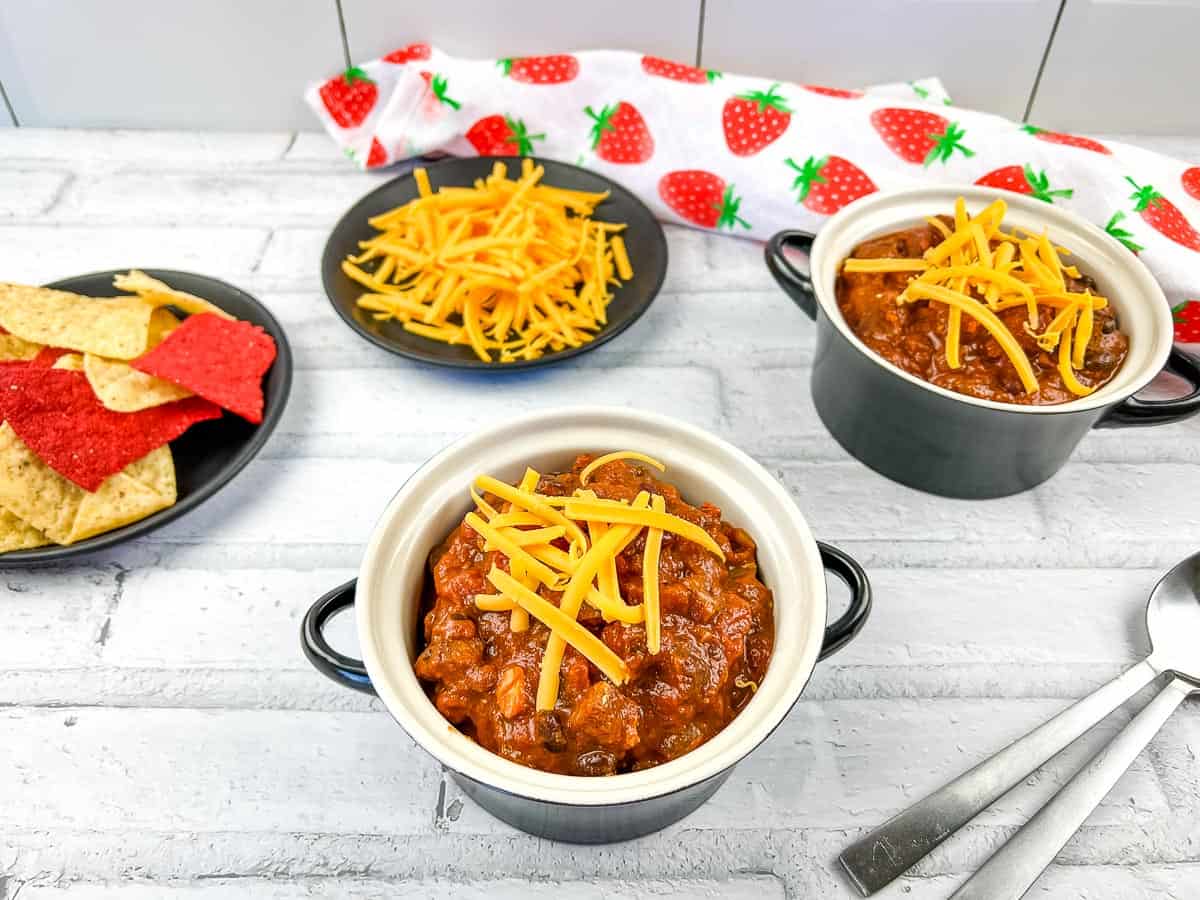 Smoked brisket chili served in two black bowls topped with sour cream, surrounded by fresh tomatoes, shredded cheese, crusty bread, and two spoons on a gray surface.