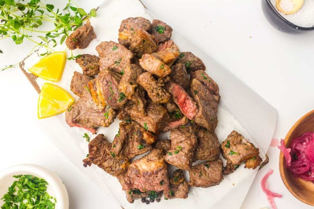 A pile of cooked, sliced beef pieces garnished with herbs on a white platter, with lemon wedges and small bowls of seasonings on the side.