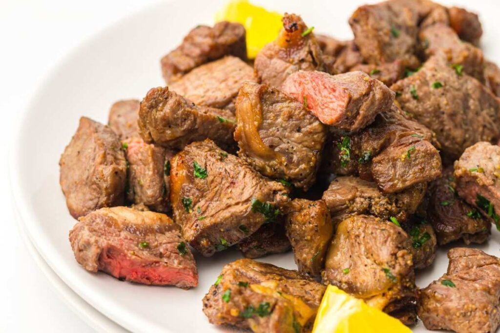 A plate of cooked steak bites garnished with herbs, with a few pieces of yellow bell pepper on the side.