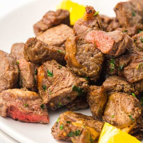 A plate of cooked steak bites garnished with herbs, with a few pieces of yellow bell pepper on the side.