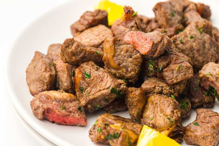 A plate of cooked steak bites garnished with herbs, with a few pieces of yellow bell pepper on the side.