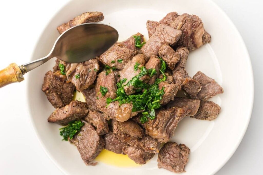 A white plate with cooked beef cubes topped with chopped herbs and olive oil, with a spoon held above the food.