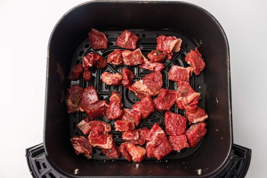 Cubed pieces of raw, seasoned beef placed inside the basket of an air fryer, ready for cooking.