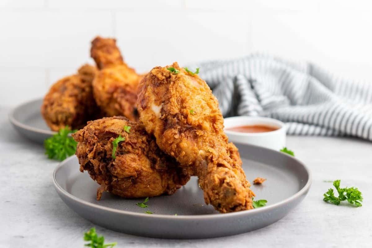 A plate with pieces of fried chicken garnished with parsley, with a bowl of sauce and a striped cloth in the background.