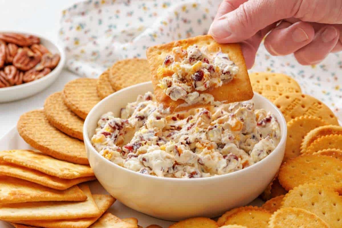 A hand dips a cracker into a bowl of cranberry cheese spread, surrounded by assorted crackers on a plate.