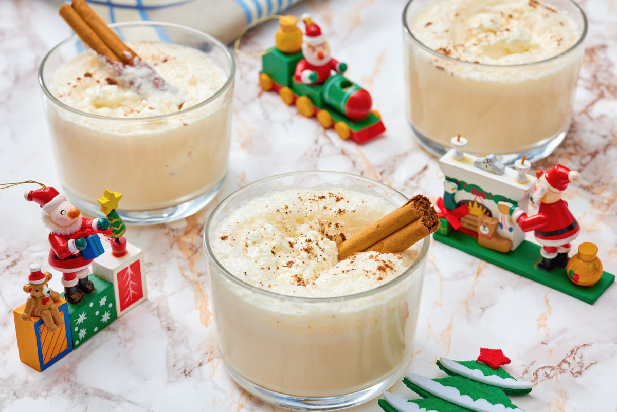 Three glasses of eggnog with cinnamon sticks on a marble surface, surrounded by small Christmas-themed figurines and decorations.