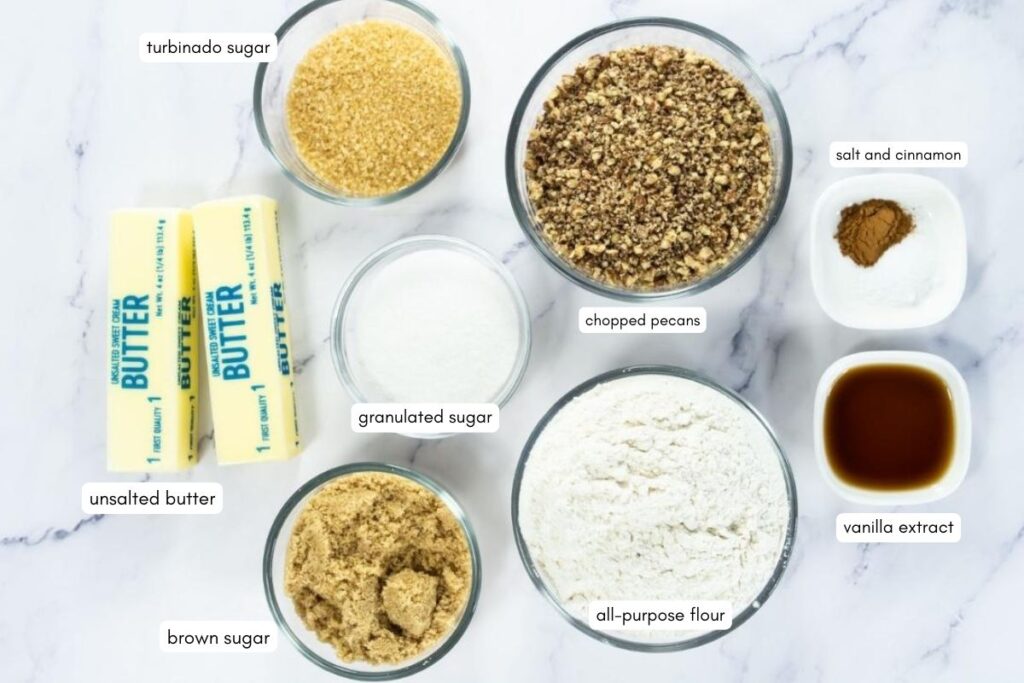Ingredients for baking displayed on a white surface: turbinado sugar, chopped pecans, granulated sugar, salt and cinnamon, unsalted butter, brown sugar, all-purpose flour, and vanilla extract.