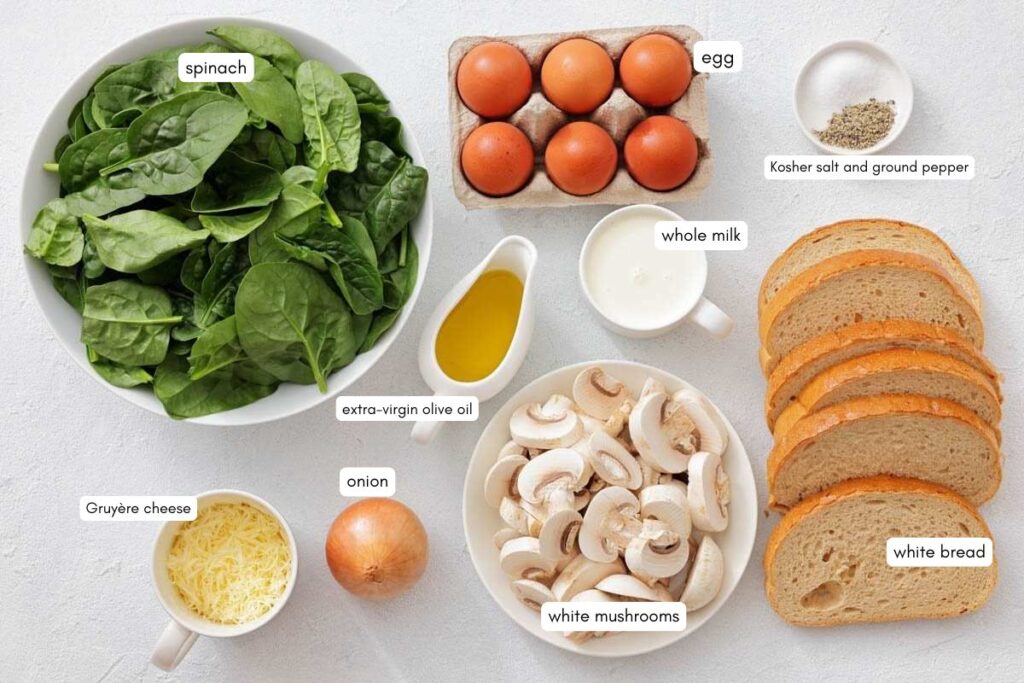 Various ingredients for a recipe, including spinach, eggs, whole milk, white bread, mushrooms, onion, Gruy&egrave;re cheese, olive oil, and a small bowl of salt and pepper.