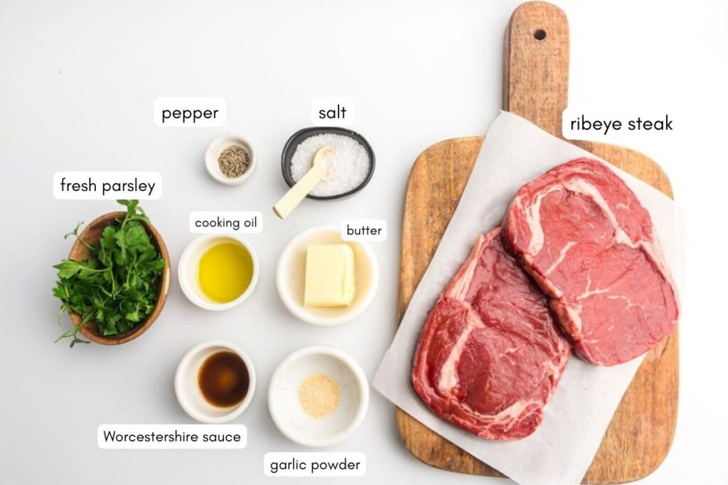 Ribeye steaks on a cutting board with bowls of fresh parsley, pepper, salt, cooking oil, butter, Worcestershire sauce, and garlic powder arranged nearby.