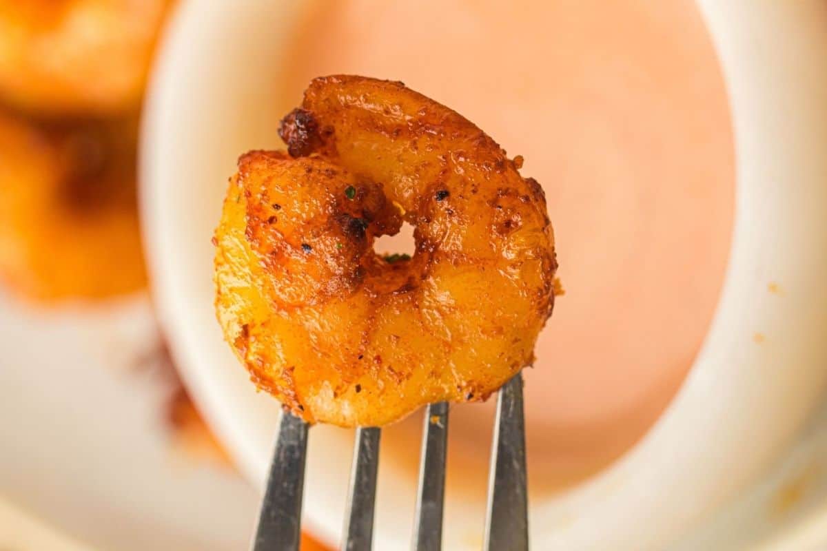 A close-up of a seasoned, cooked shrimp on a fork, held in front of a bowl of dipping sauce.