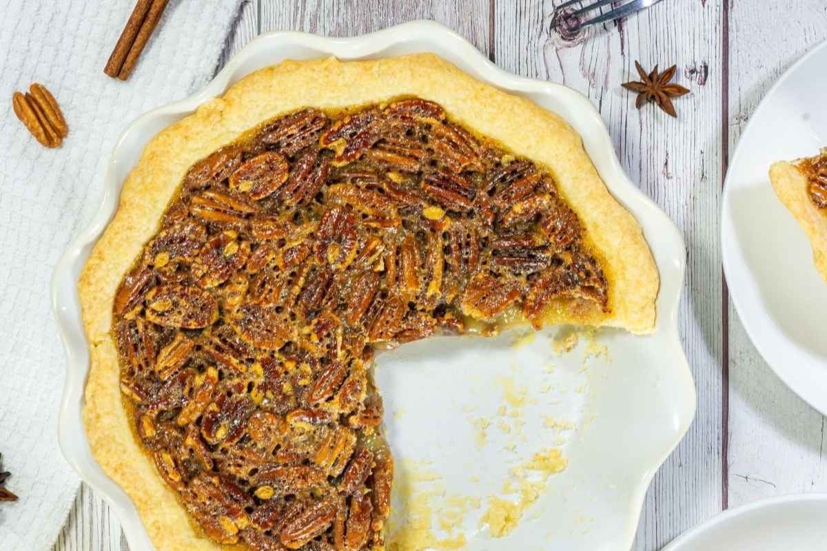 A pecan pie in a white pie dish with a large slice missing, displayed on a light wooden surface.