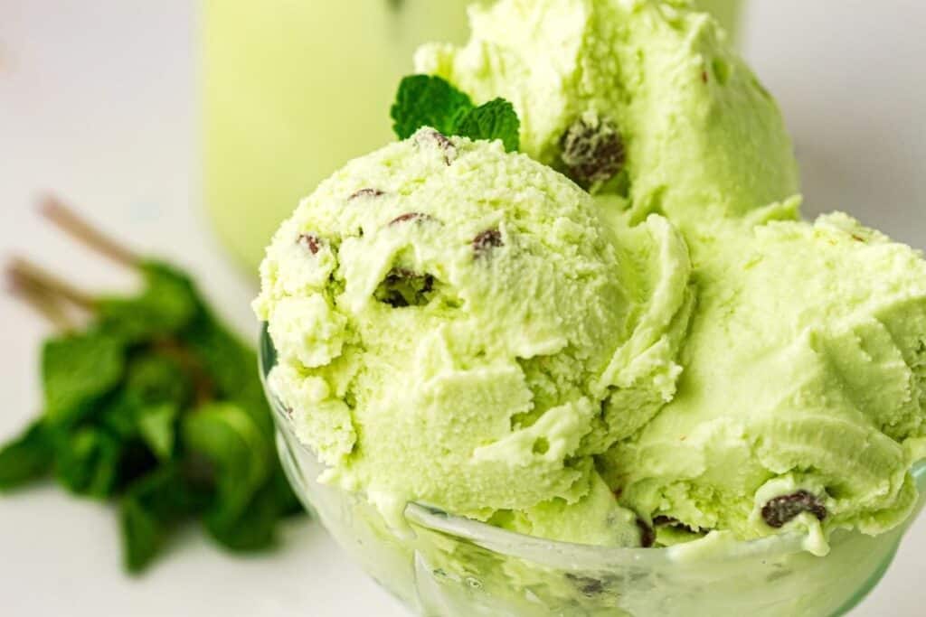 Three scoops of mint chocolate chip ice cream in a glass bowl, with chocolate pieces visible and a sprig of mint in the background.