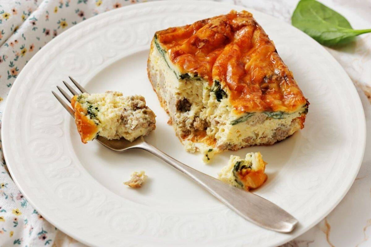 A slice of Sausage, Spinach, and egg casserole on a white plate, with a fork holding a bite-sized piece.