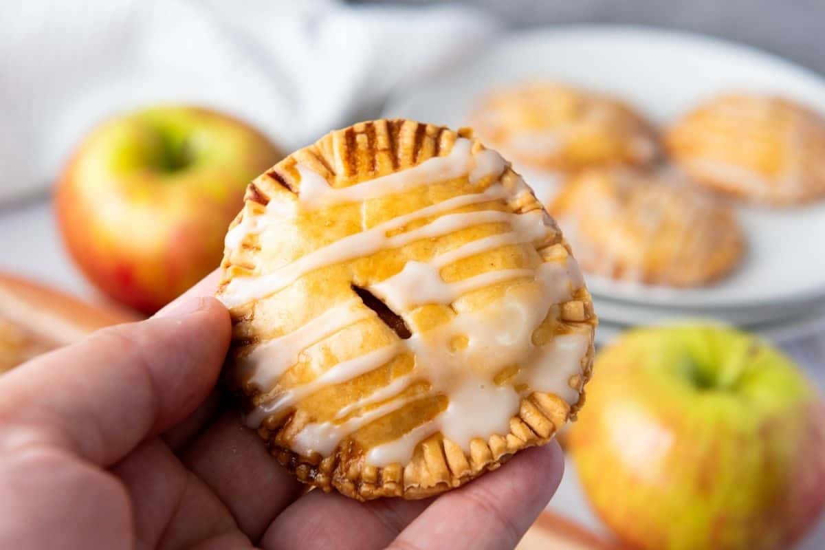 A hand holds a small round hand pie with icing drizzle; apples and more hand pies are visible in the background.