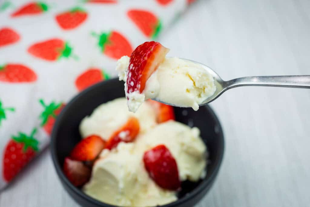 A spoonful of vanilla ice cream and a strawberry held above a bowl with more ice cream and strawberries, next to a strawberry-patterned cloth.