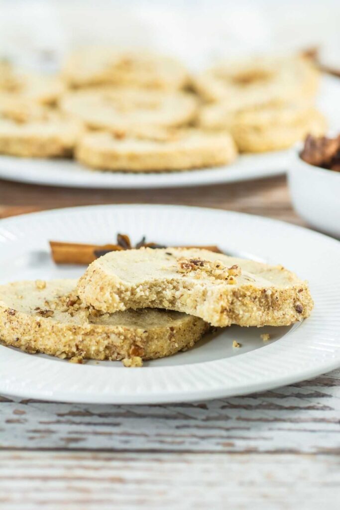 Close-up of two round shortbread cookies, one partially eaten, on a white plate with more cookies and a bowl in the background.