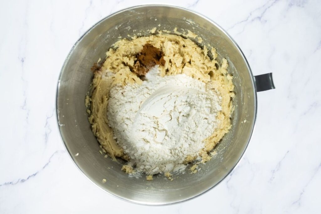 A metal mixing bowl with cookie dough, flour, and brown sugar on top, resting on a white marble surface.