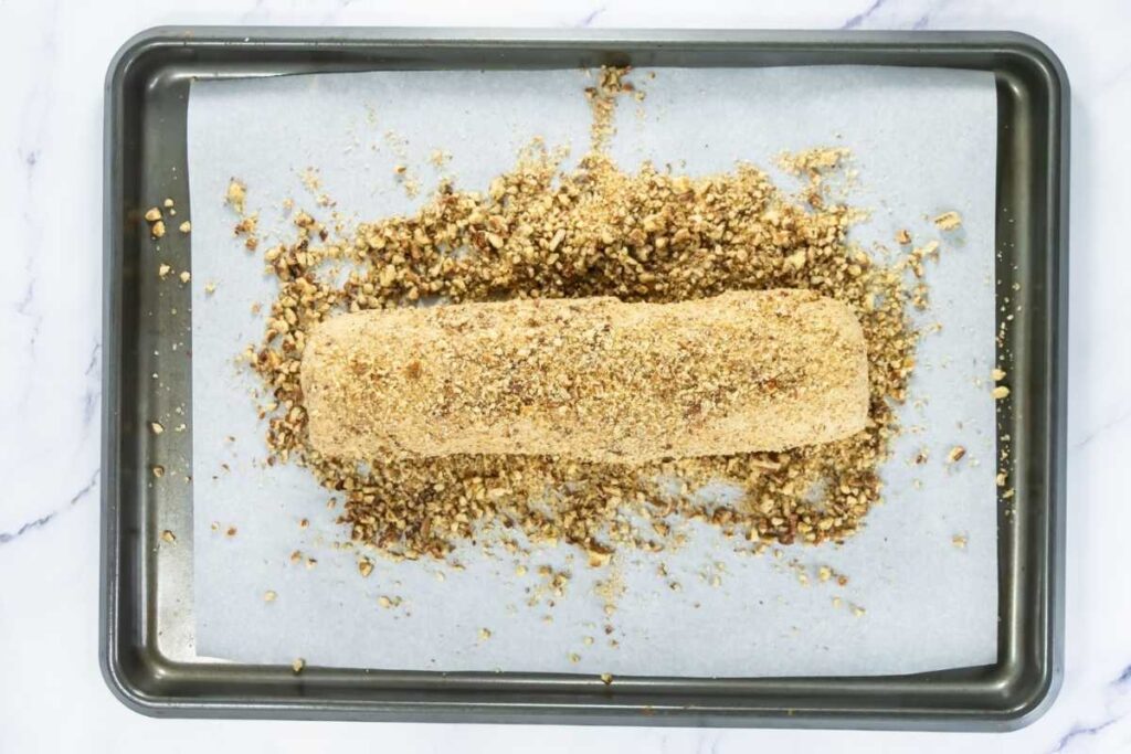 A pastry log coated in crushed nuts and crumbs sits on parchment paper on a baking tray.