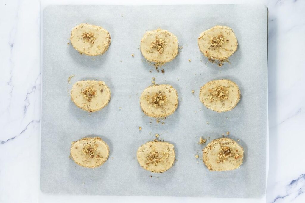 Nine unbaked cookie dough rounds topped with pecans are arranged on a parchment-lined baking sheet.