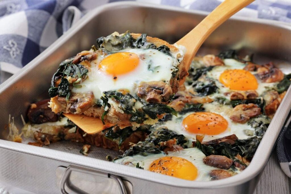 A baking dish with baked eggs, sautéed spinach, mushrooms, and onions, with one serving being lifted out on a spatula.