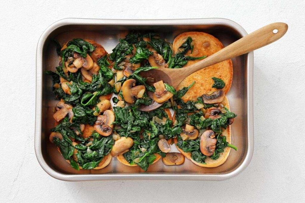 A metal baking dish containing saut&eacute;ed spinach and mushrooms served over slices of toasted bread, with a wooden spoon resting inside.