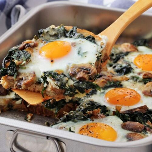 A baking dish with baked eggs, saut&eacute;ed spinach, mushrooms, and onions, with one serving being lifted out on a spatula.