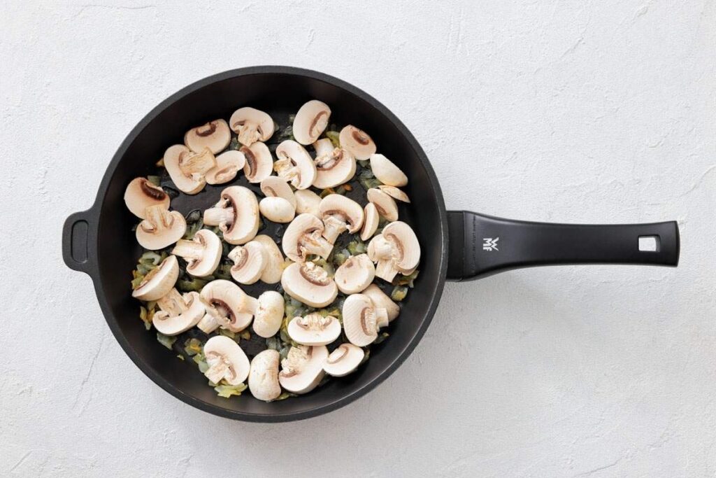 A black frying pan with sliced mushrooms and onions being saut&eacute;ed on a light-colored surface.