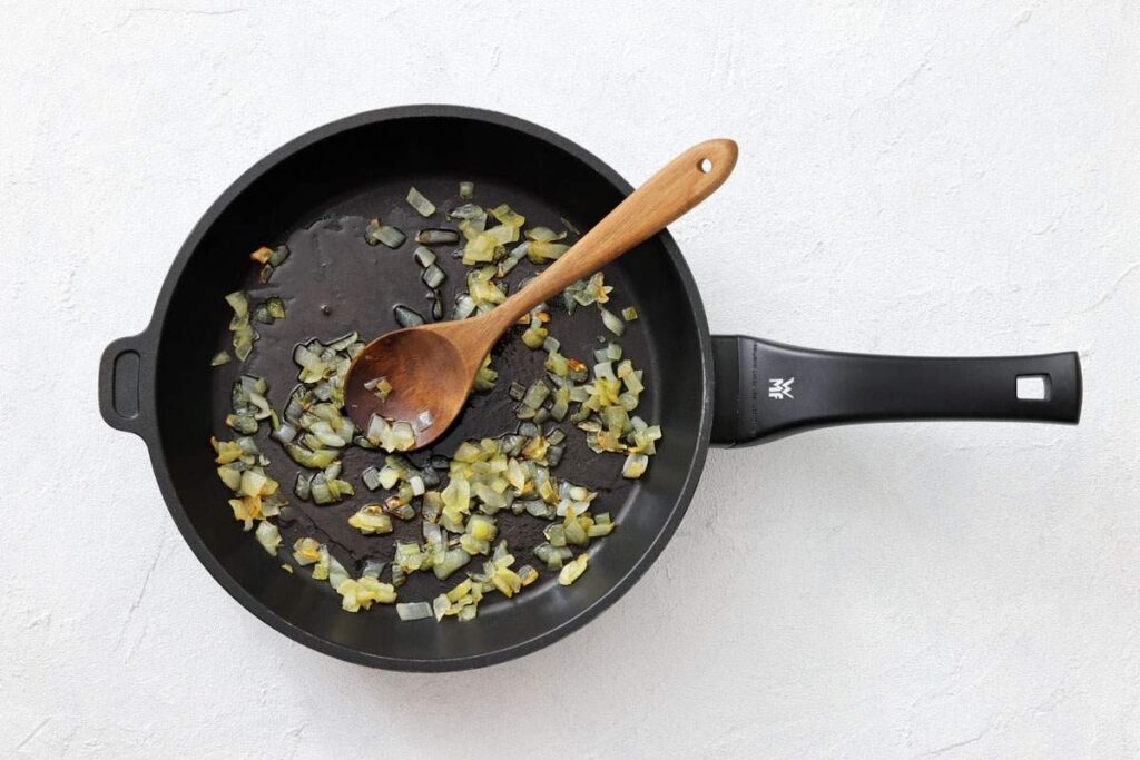 A black frying pan with saut&eacute;ed diced onions and a wooden spoon on a light-colored surface.