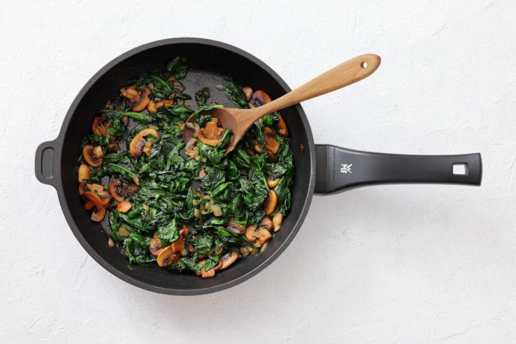 A black frying pan with cooked spinach and sliced mushrooms, stirred with a wooden spoon, sits on a white surface.